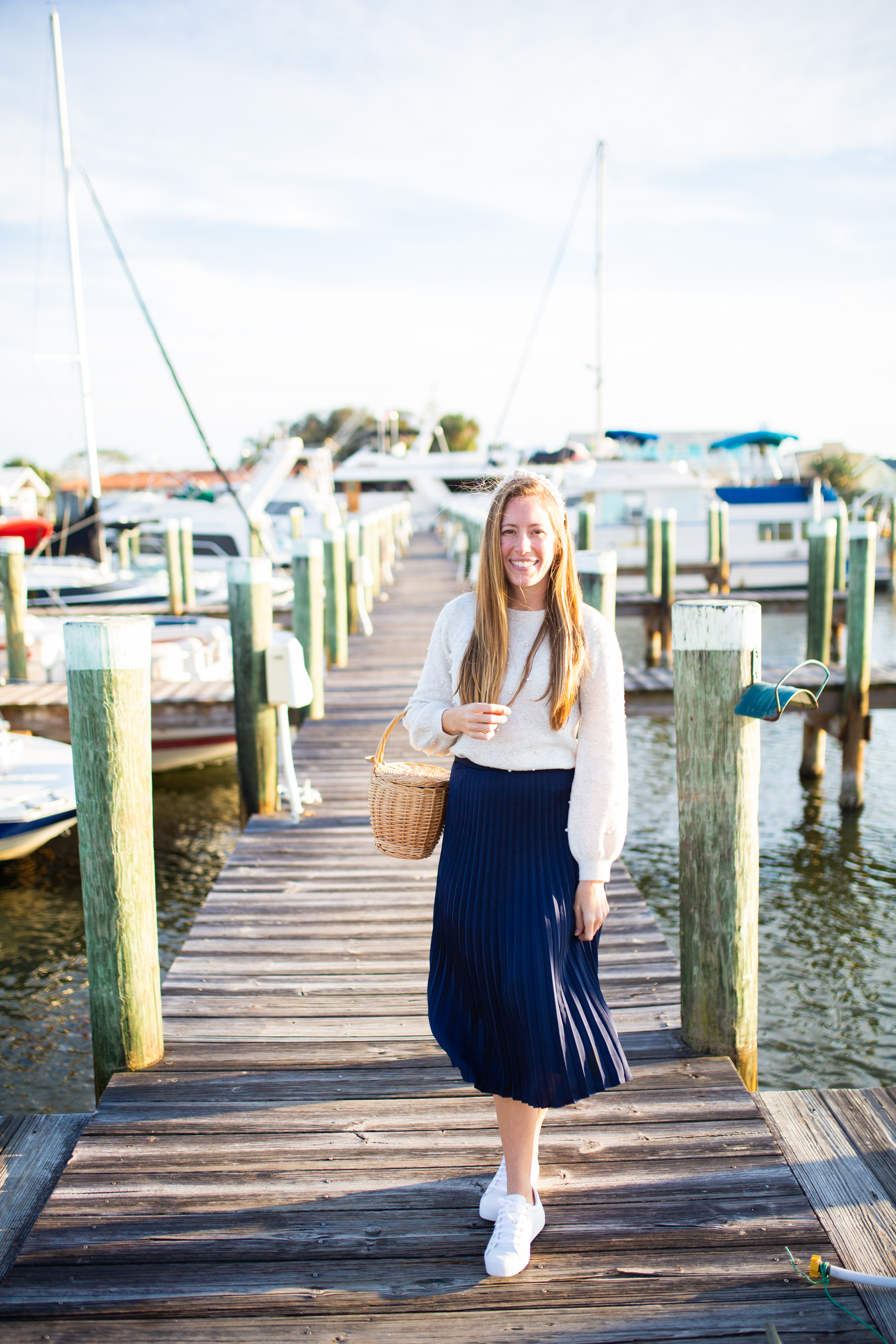 woman smiling and wearing white top, blue, skirt, and white sneakers at the docks in sunshine, florida
