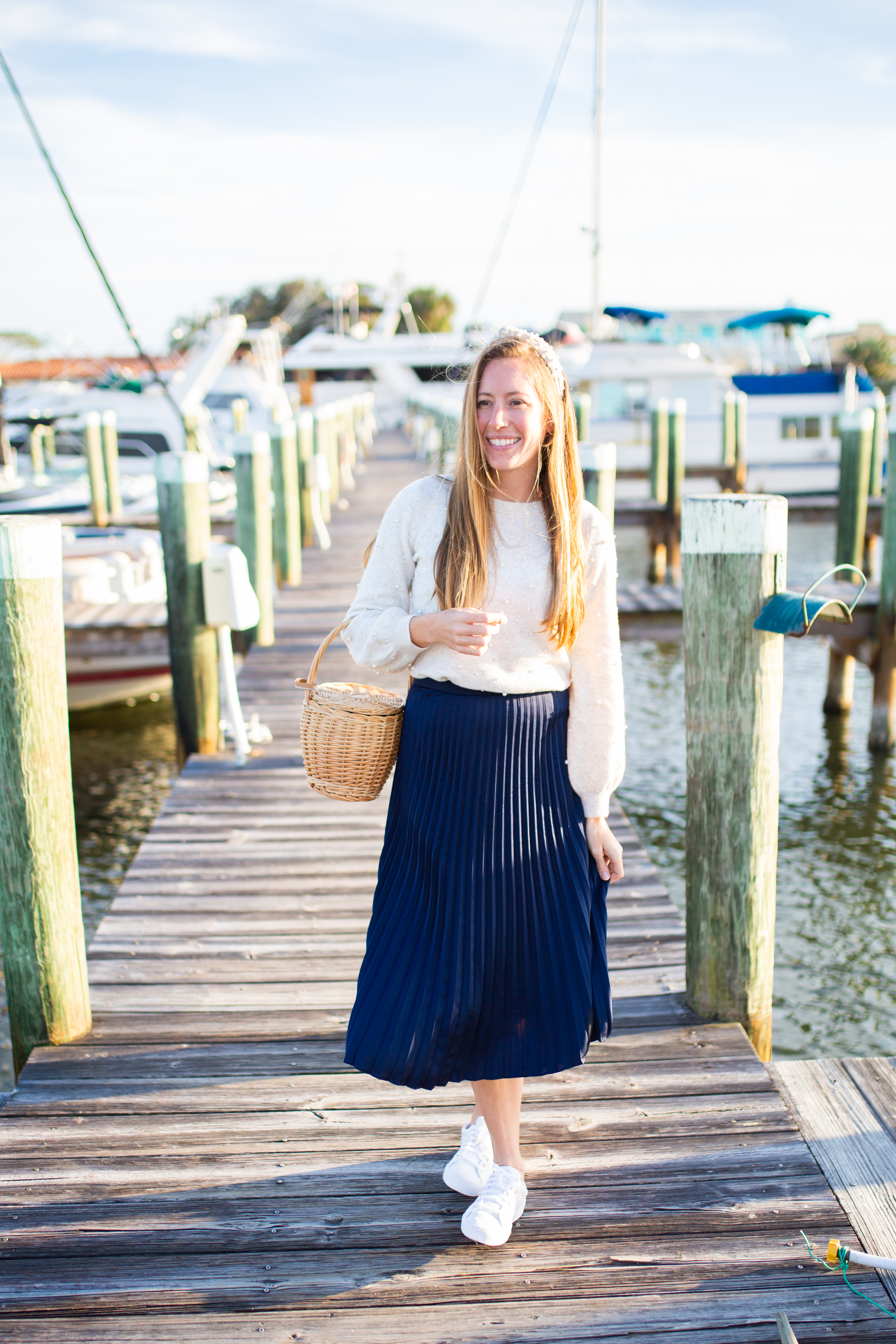 woman wearing white Sweaters for Spring, blue, skirt, and white sneakers at the docks in sunshine, florida