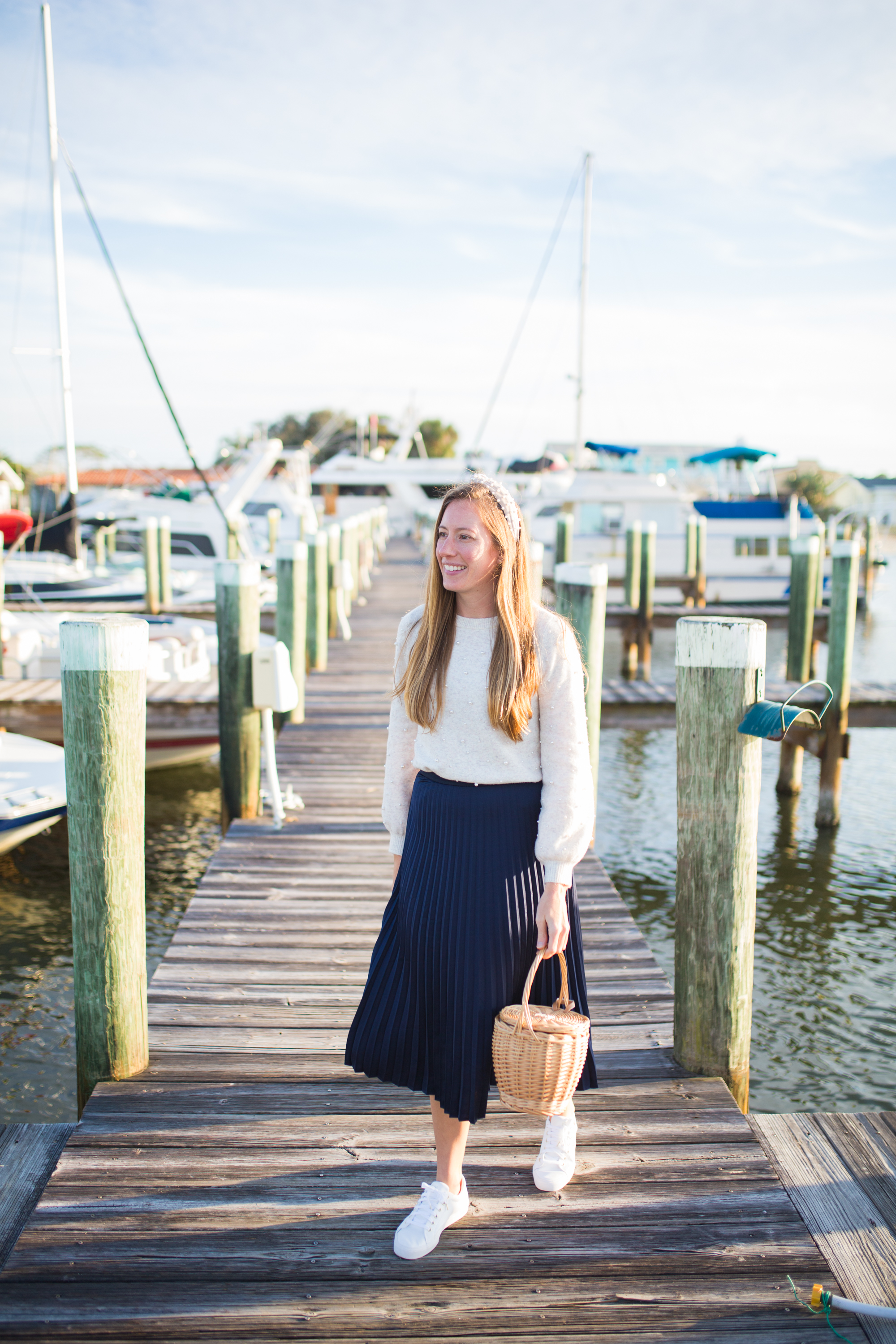 woman wearing white Sweaters for Spring, blue, skirt, and white sneakers and holding a weaved bag at the docks in sunshine, florida