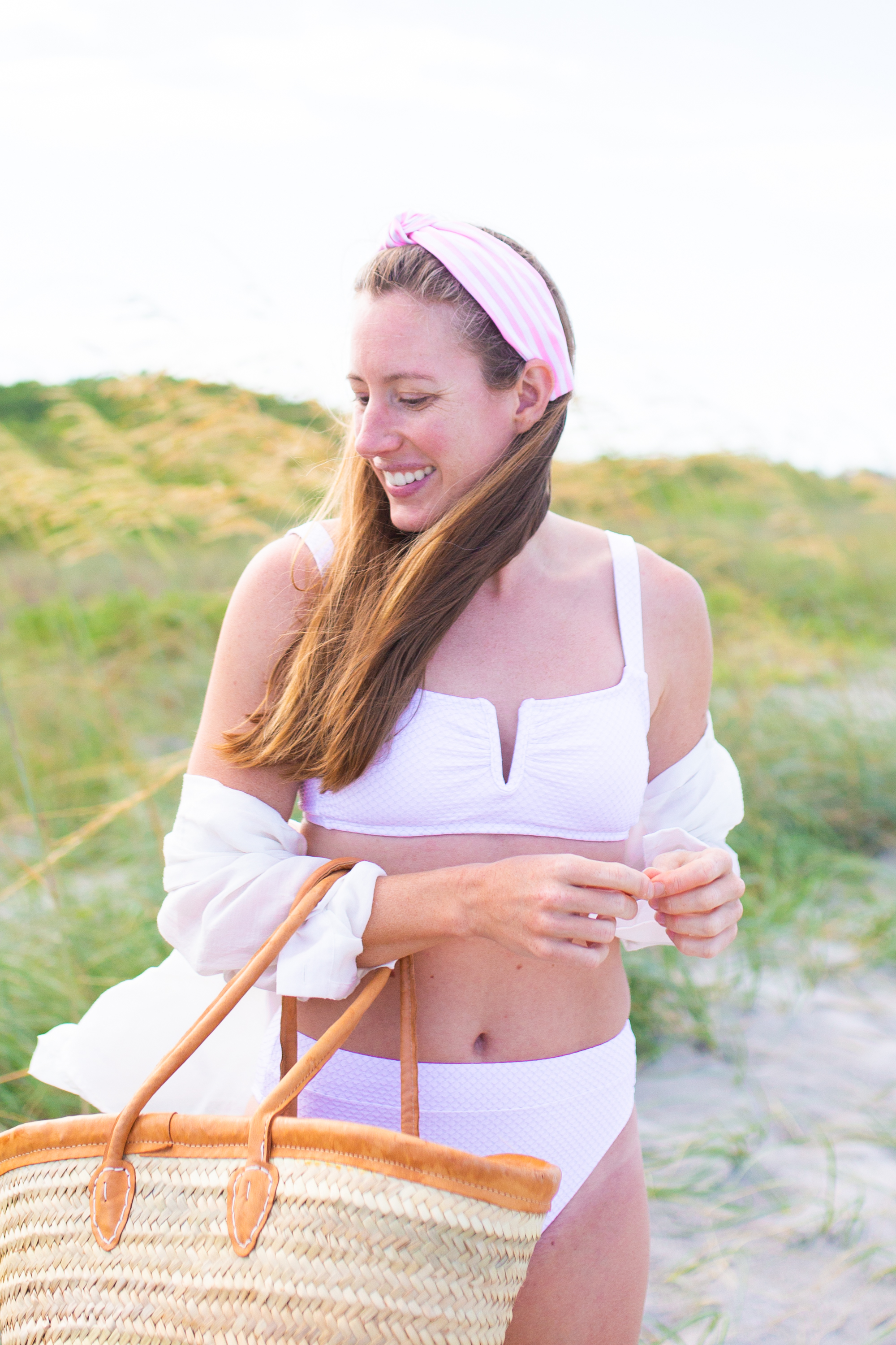 woman smiling at the beach and wearing a two piece Swimsuit, wearing a pink headband, and holding weaved bag