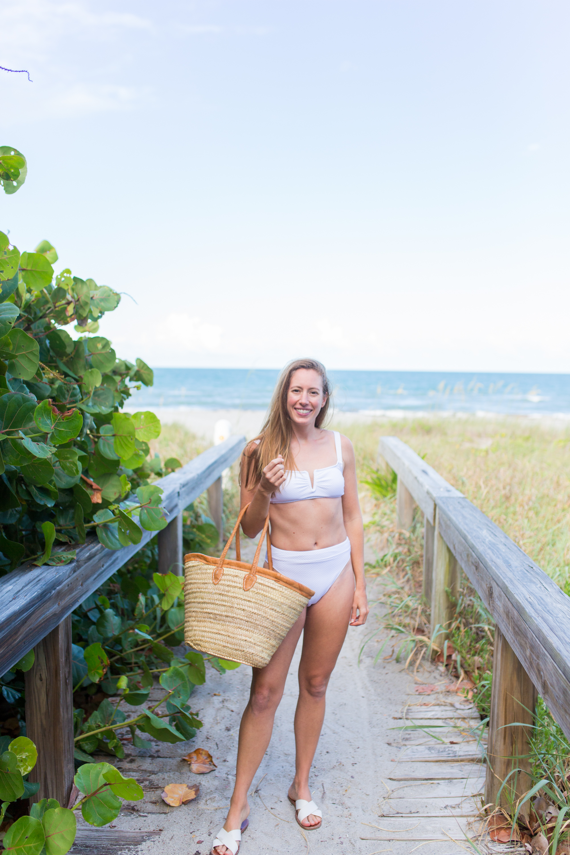 woman at the beach and wearing Classic White Swimsuits, holding a weaved bag, and white flat sandals