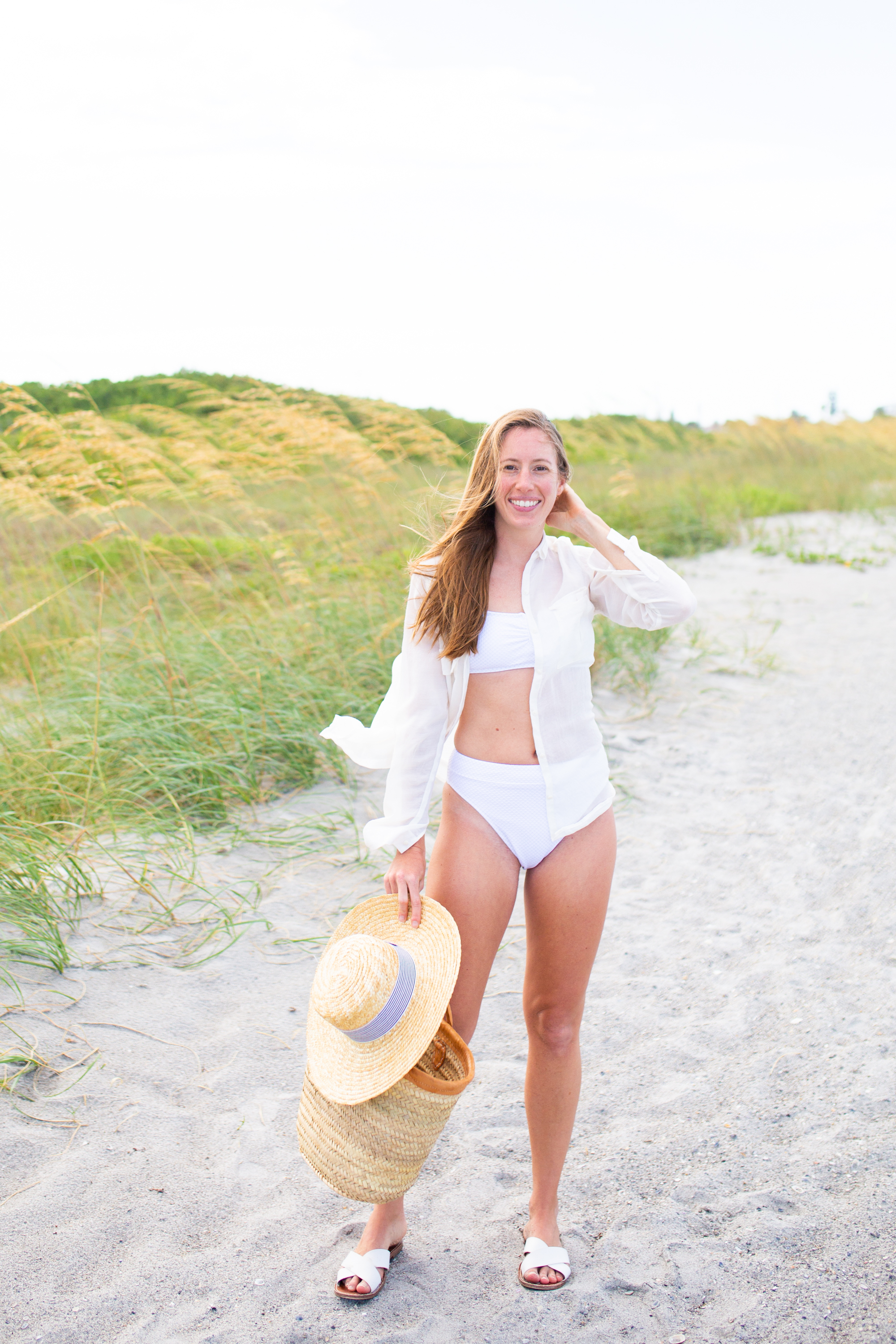 woman smiling at the beach and wearing Classic White Swimsuits, wearing a pink headband, and holding weaved bag and hat