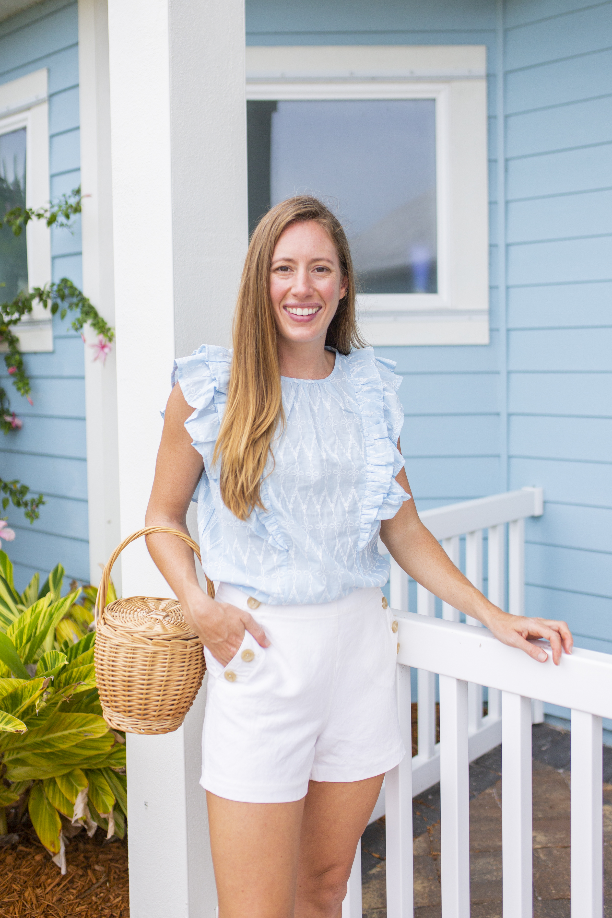 Woman smiling and wearing blue and white top styled with white shorts and a raffia bag 