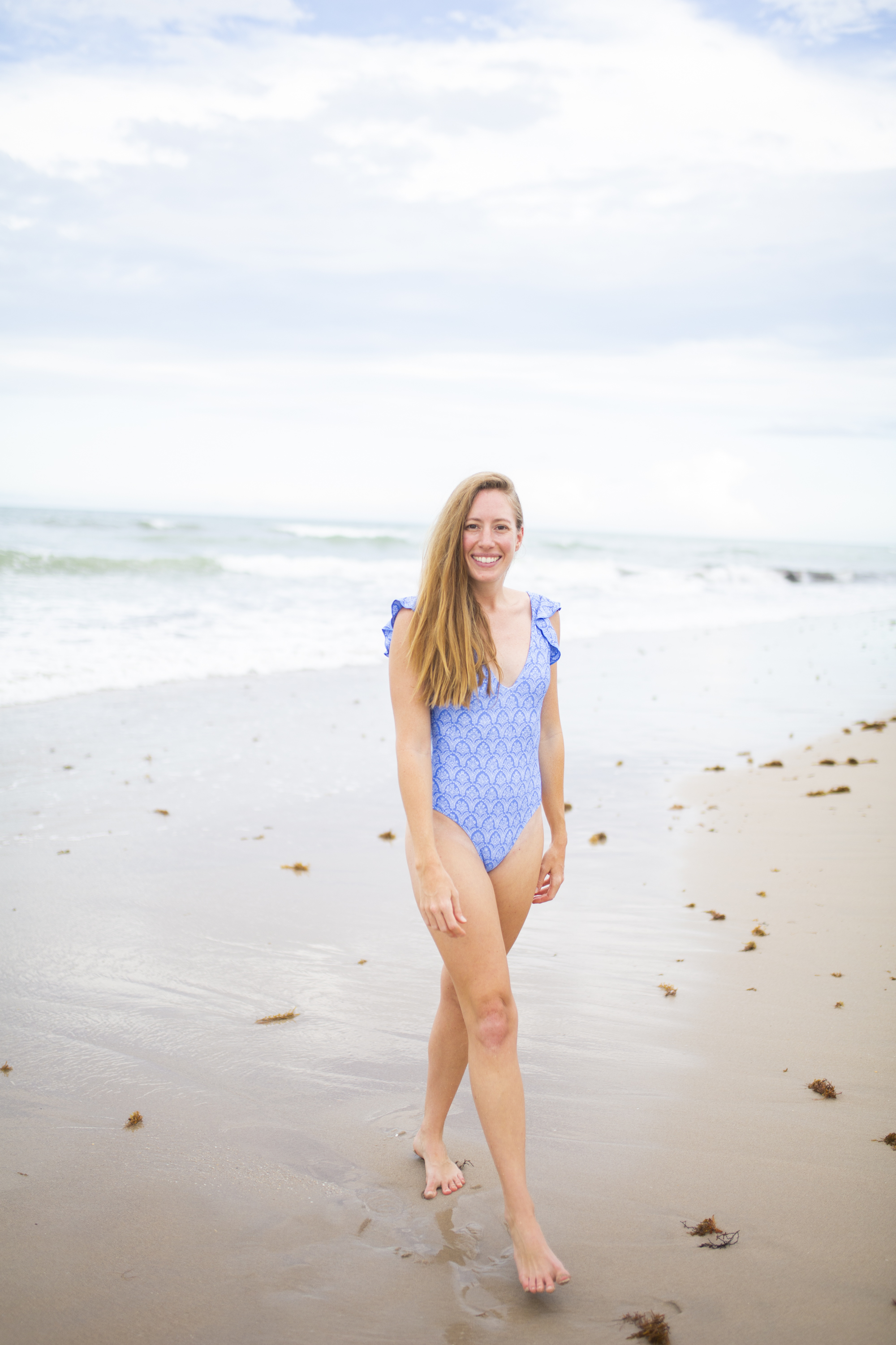 woman walking on the beach in a blue and white Summersalt one piece swimsuit 