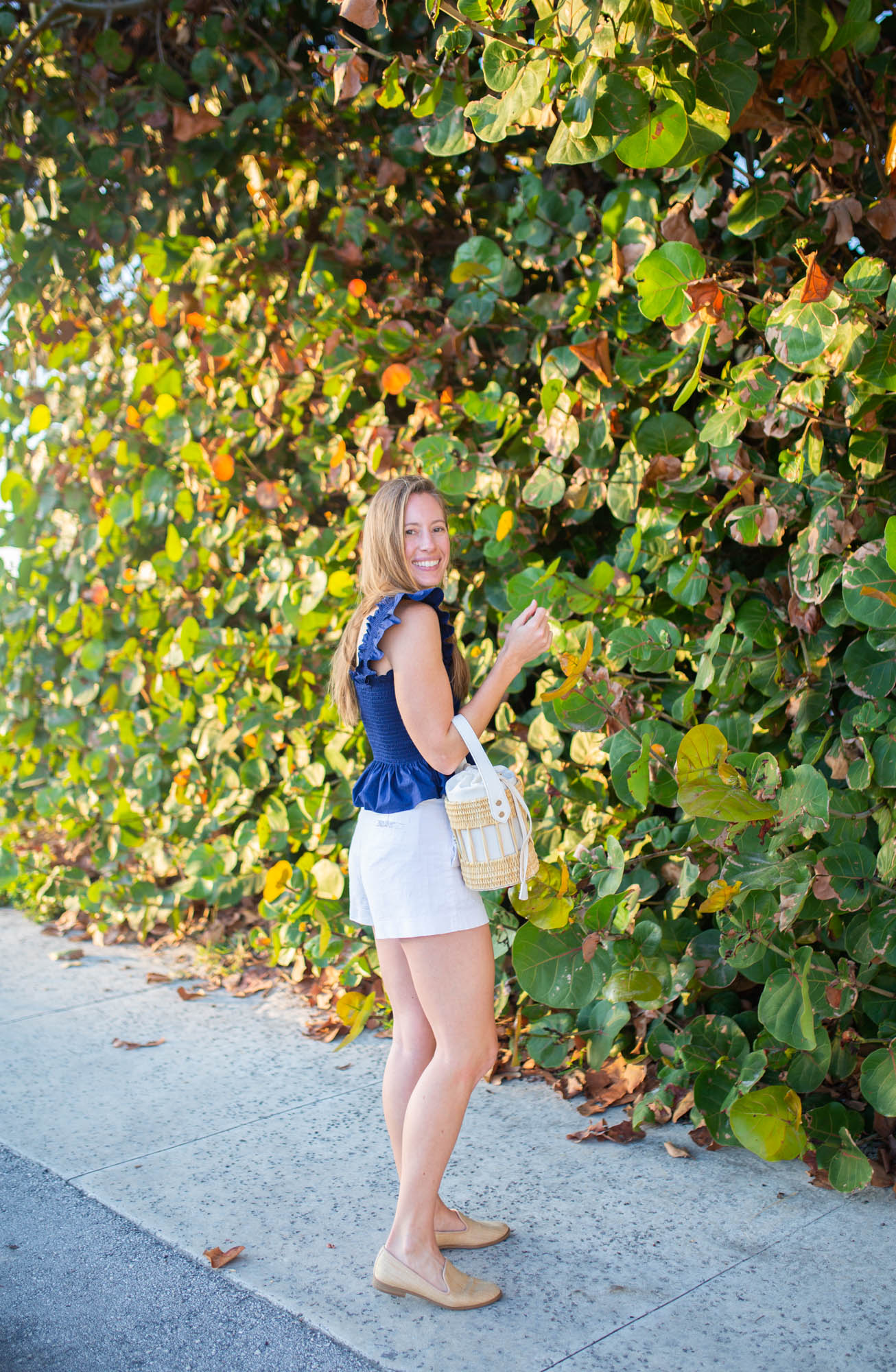 Women standing in front of sea grass wearing a navy ruffle sleeve top with white shorts, a basket bag and loafers for a beach outfit 