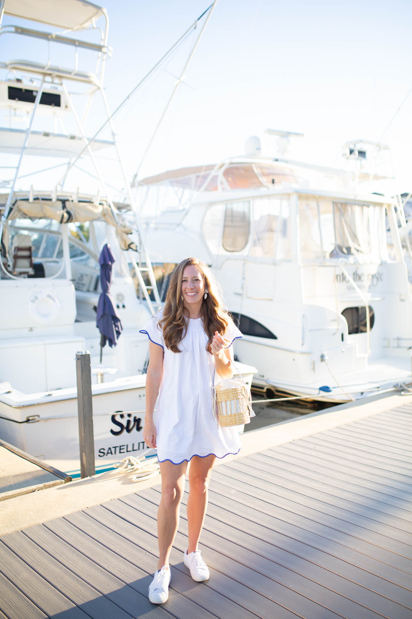 A girl wearing a casual white dress with scallop details in front of yacht boats in a marina. 