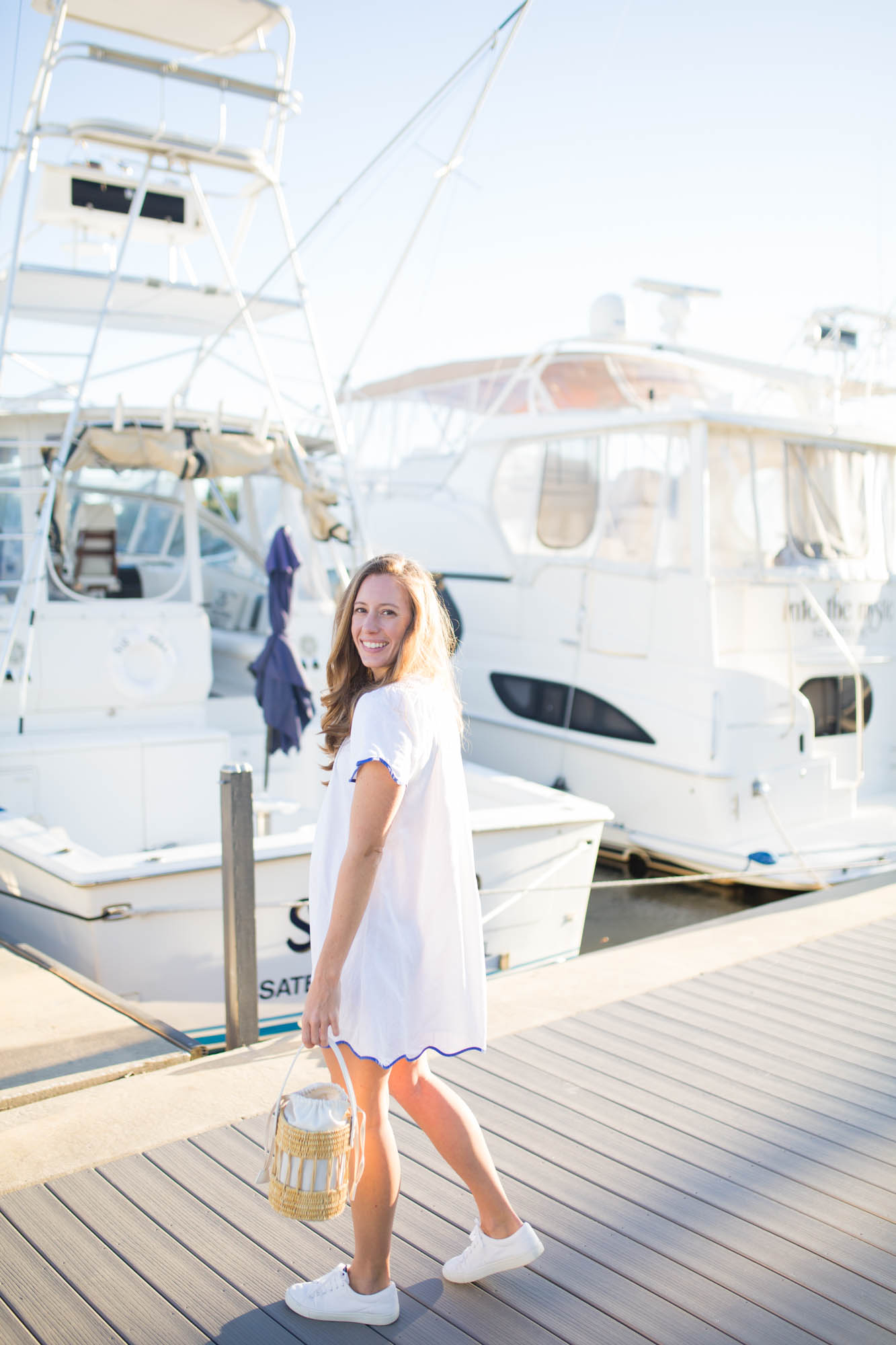 A girl wearing a casual white dress with scallop details in front of yacht boats in a marina. 