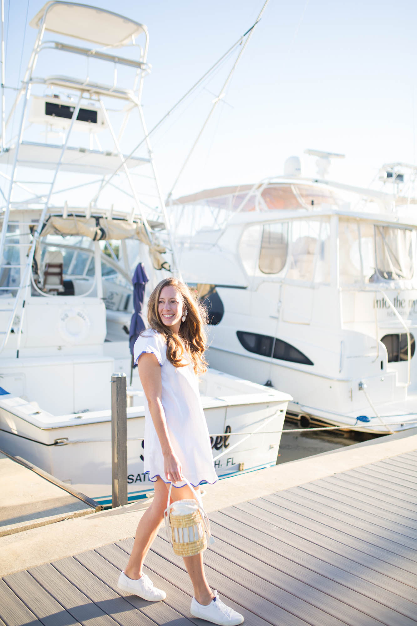 A girl wearing a casual white dress with scallop details in front of yacht boats in a marina. 